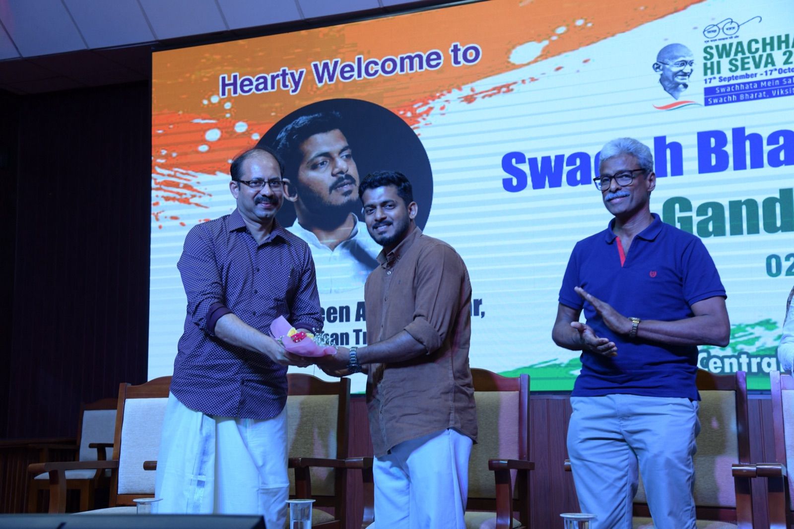 On stage, a man in a purple checked shirt hands a small bouquet to a man in a brown shirt while a third man applauds, in front of a welcome screen for the Swachh Bharath programme.