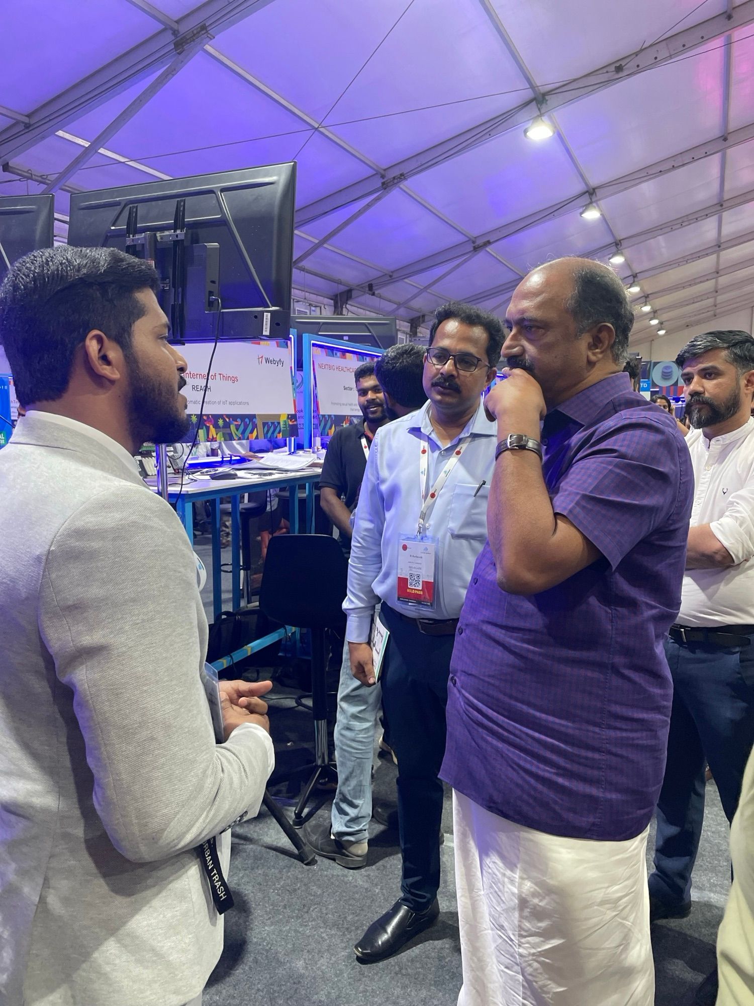 A man in a light jacket talks with a man in a purple shirt and two others inside a large tent at a startup expo.