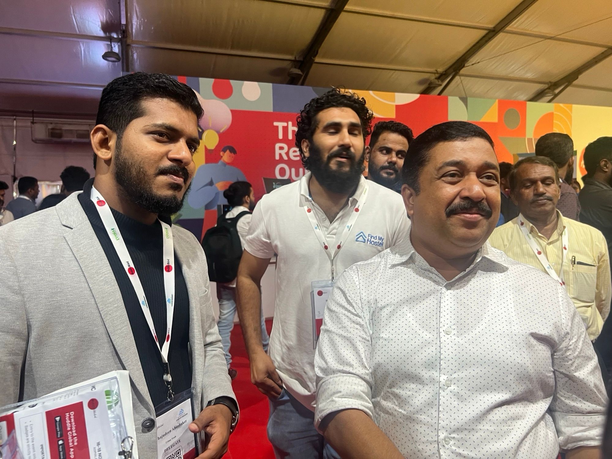 A man in a grey jacket with a name badge stands beside a smiling man in a white patterned shirt at a startup expo.