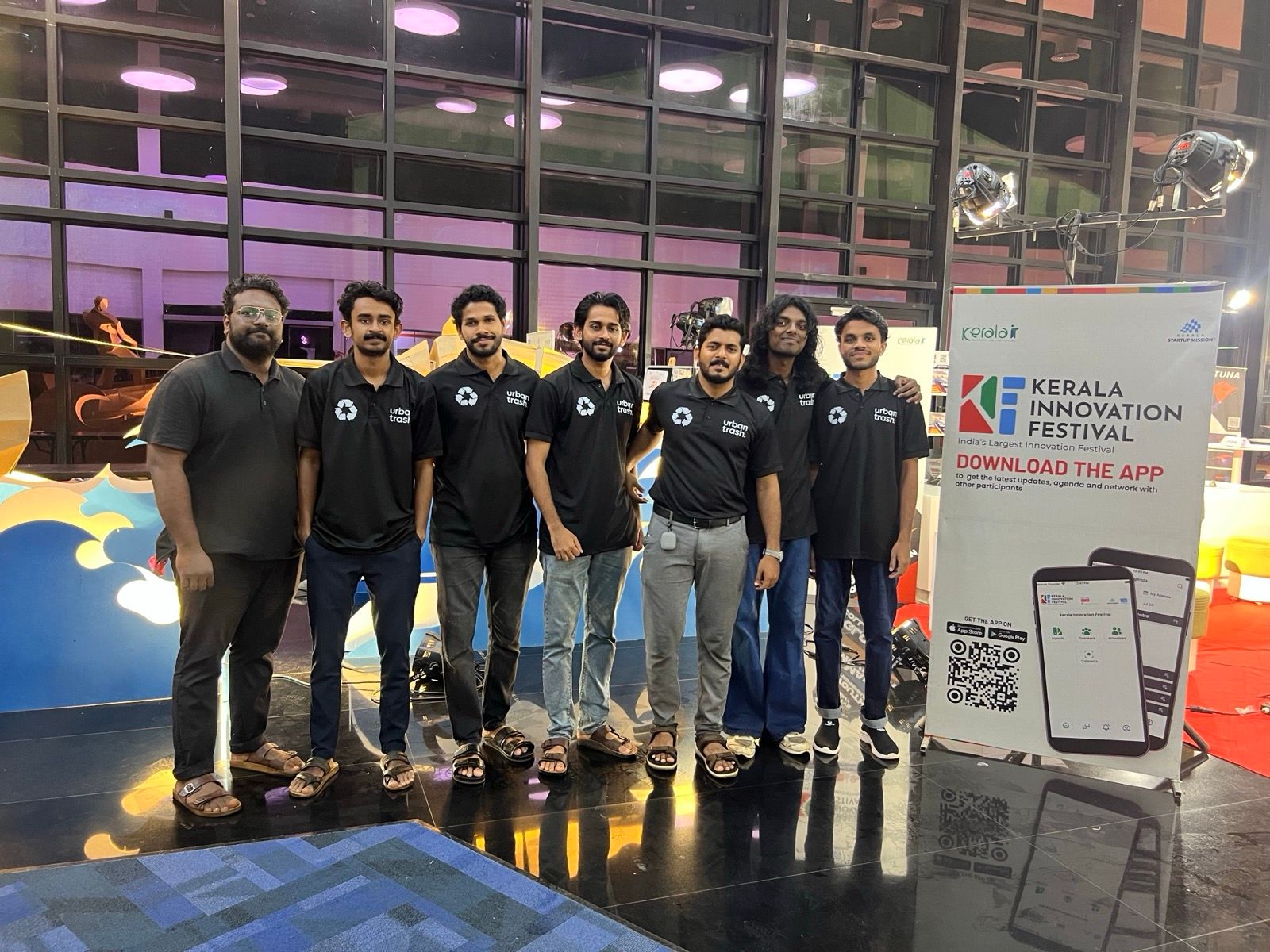 Seven people in black Urban Trash polo shirts stand in a row inside a glass-walled hall, beside a Kerala Innovation Festival banner.