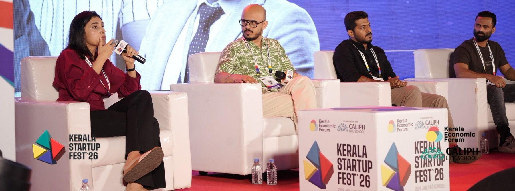 Four panelists in white armchairs on stage at Kerala Startup Fest 2026. A woman on the left speaks into a microphone while the others listen.