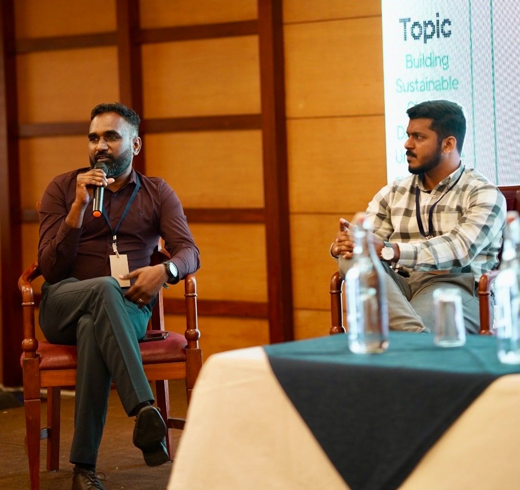 Two men seated in wooden chairs on a panel stage, one speaking into a microphone, with a slide on sustainability behind them.