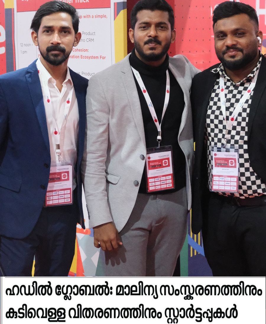 A Malayalam newspaper clipping. A photo of three men standing close together in conference lanyards, in front of colourful event signs, sits above a headline about Huddle Global and startups for waste processing and drinking-water distribution.