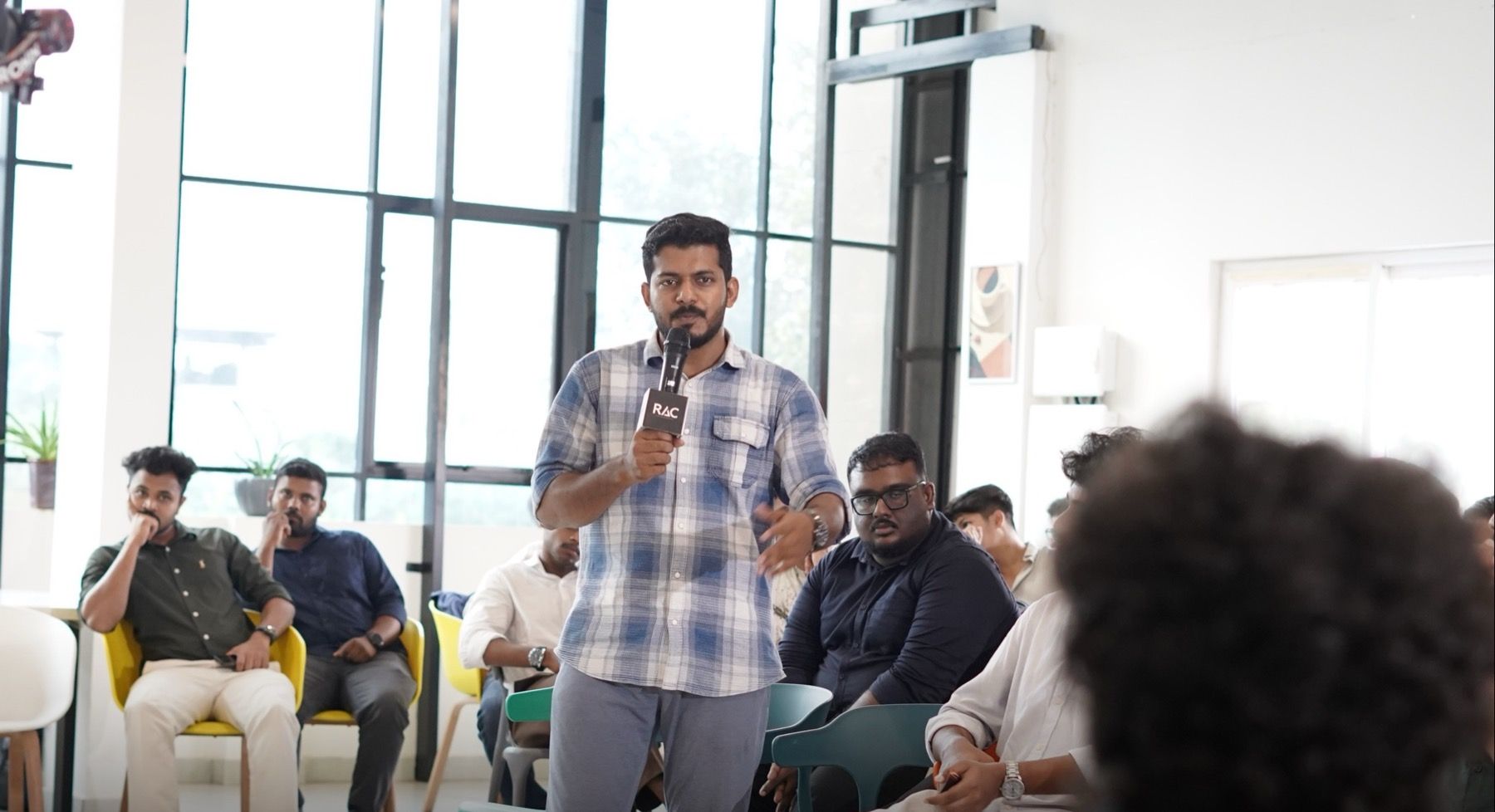 A man in a blue check shirt stands and speaks into a microphone in a bright room, with an audience seated around him.