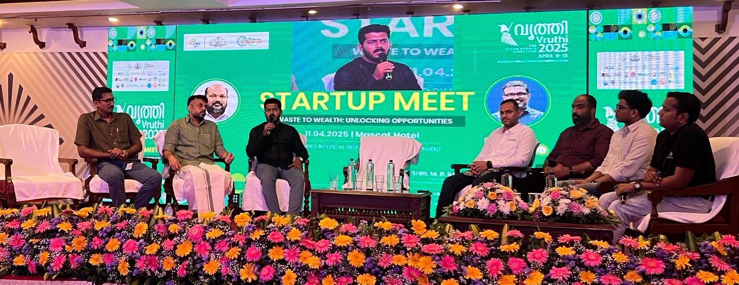 Seven people on a flower-edged stage under a green screen headed Startup Meet, Waste to Wealth. One of the panelists holds a microphone and speaks.